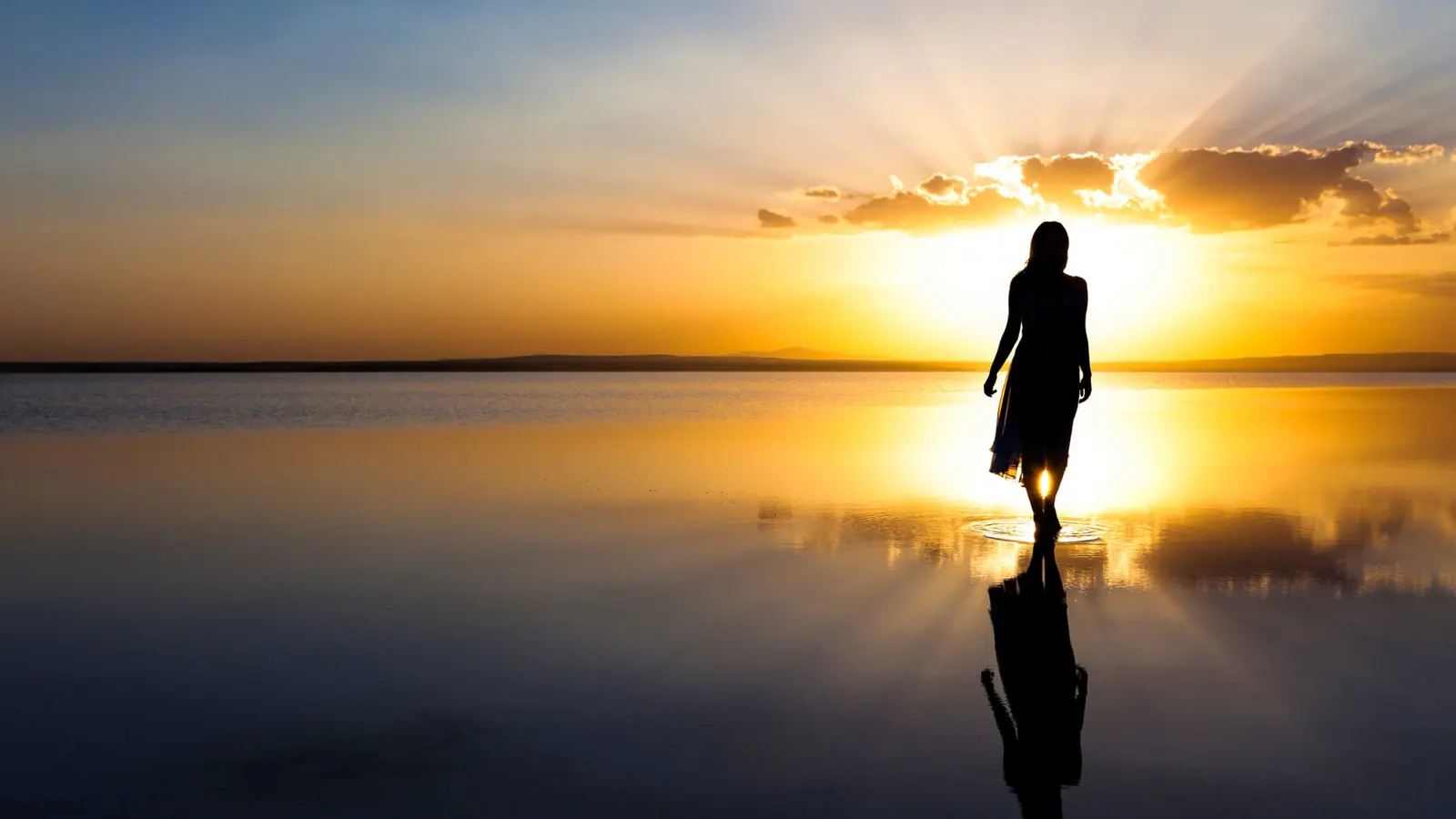 A figure standing in still water at sunrise, arms open toward the light